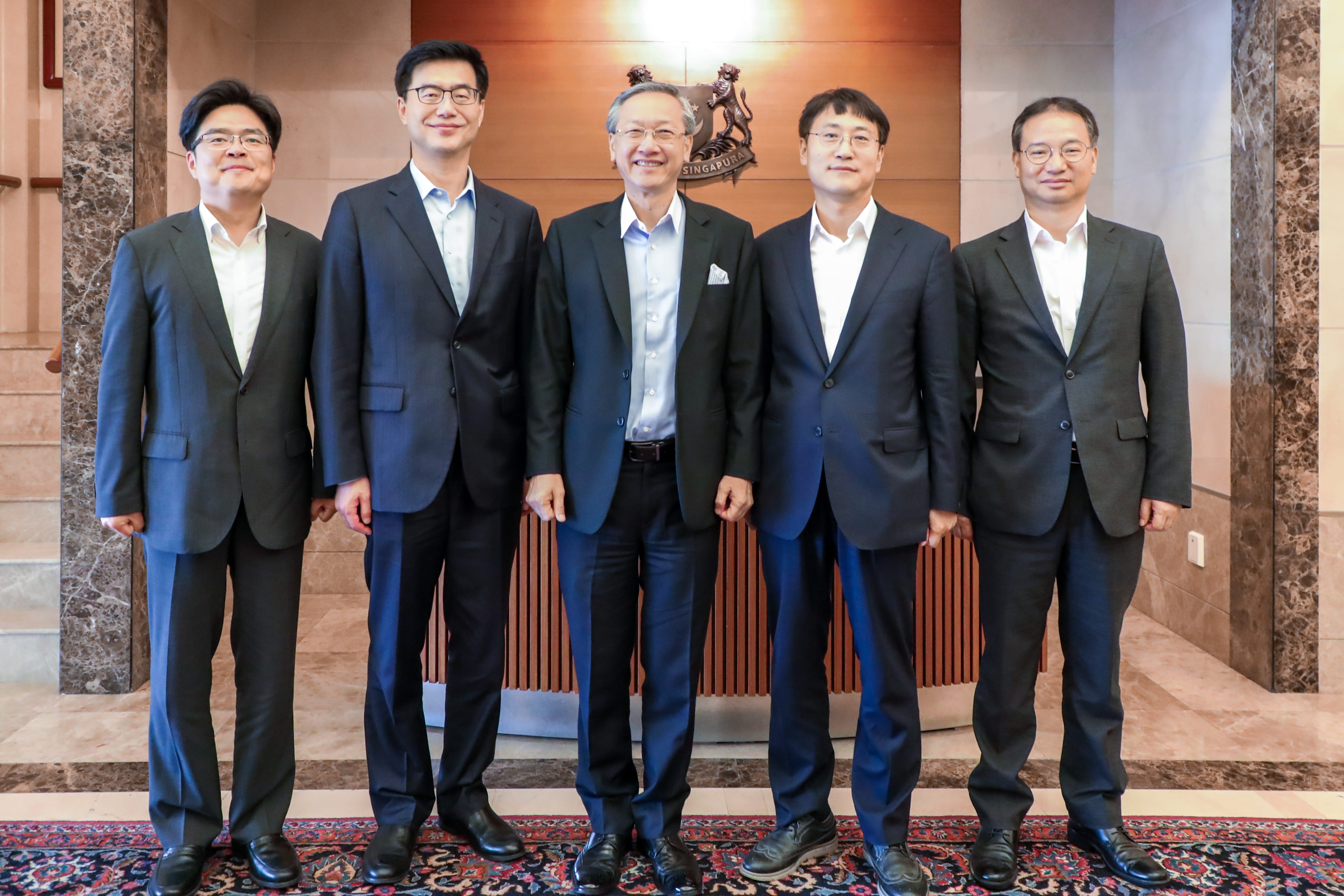 Five men in suits stand in a row before the Singapore coat of arms.